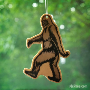 Bigfoot or sasquatch air freshener with a white string against a blurred leafy green background.