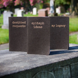 Three pocket-sized notebooks, each with a different inscription in serif font - from left to right, "deathbed requests," "epitaph ideas," and "my legacy." The notebooks are taken on top a gravestone with a blurred cemetary image in the background.