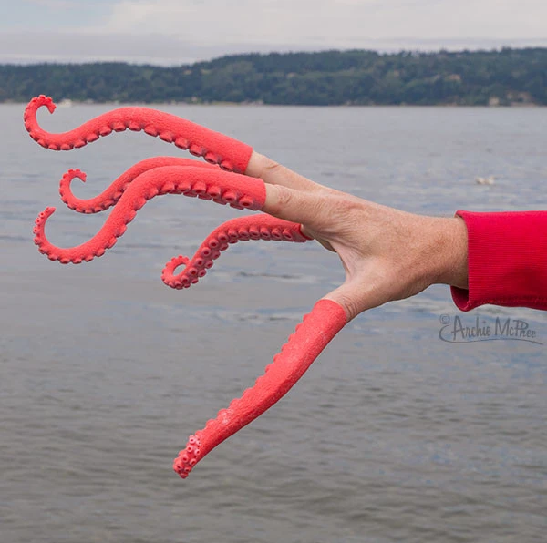 A human hand wearing five tentacle-shaped finger puppets. The tentacles are a coral red color and extend roughly four inches from the fingertips. The image backdrop is a large body of water and some land.